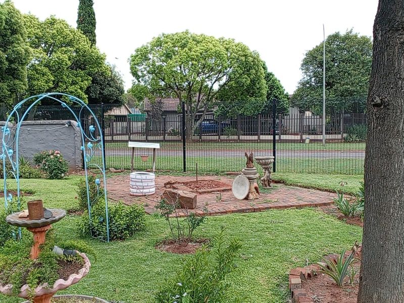 A landscaped garden featuring a brick paved area, mature trees, and metal perimeter fencing.