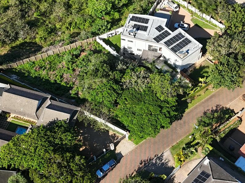 Aerial view of a residential property featuring a main dwelling with solar panels, established garden landscaping, and a paved driveway within a suburban estate.