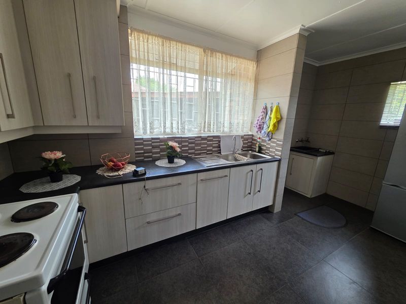 A functional kitchen featuring light wood-grain cabinetry, dark laminate countertops, tiled backsplash, and tiled flooring with a built-in sink and electric stove.