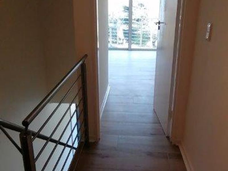 A bright hallway featuring modern tiled flooring and a metal balustrade, leading through an open doorway toward a sunlit room with sliding glass doors.