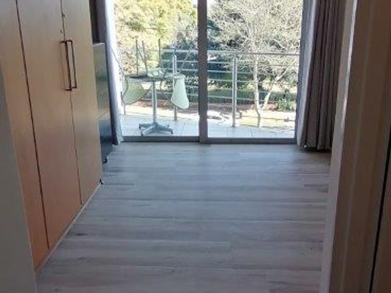 A bright, empty bedroom featuring modern laminate flooring, built-in cupboards, and a large sliding glass door providing access to a private balcony.