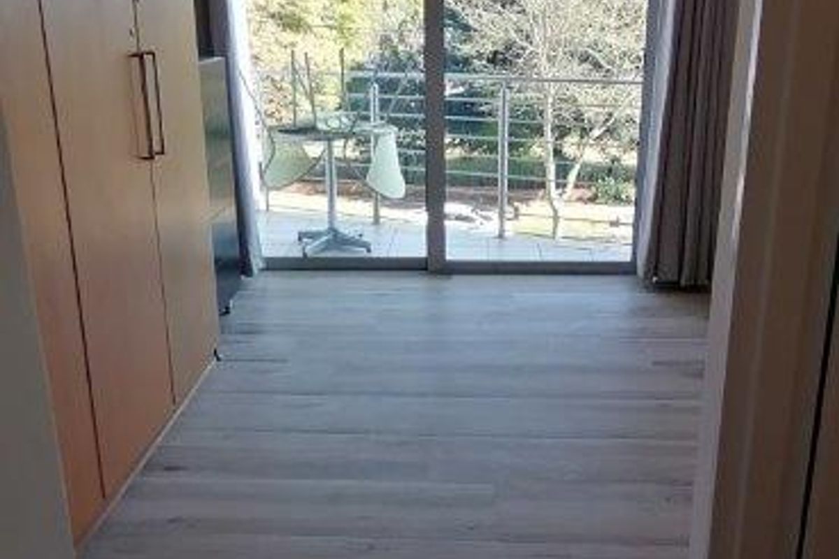 A bright, empty bedroom featuring modern laminate flooring, built-in cupboards, and a large sliding glass door providing access to a private balcony.