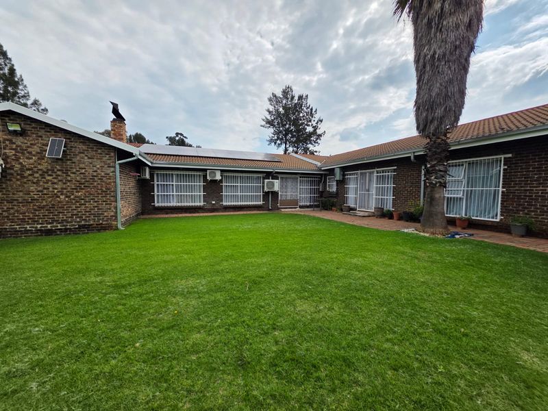 A spacious rear garden featuring a well-maintained lawn, brick-paved patio area, and a classic brick-faced house exterior with solar panels and security window bars.