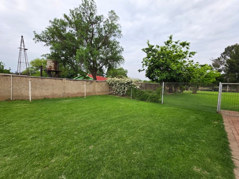 A spacious backyard featuring a well-maintained lawn, perimeter wall, wire fencing, and a rustic water tank with a windmill structure in the background.