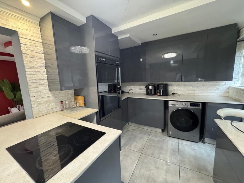 Modern kitchen featuring high-gloss grey cabinetry, stone countertops, built-in oven, electric hob, and stone-clad backsplash with tiled flooring.