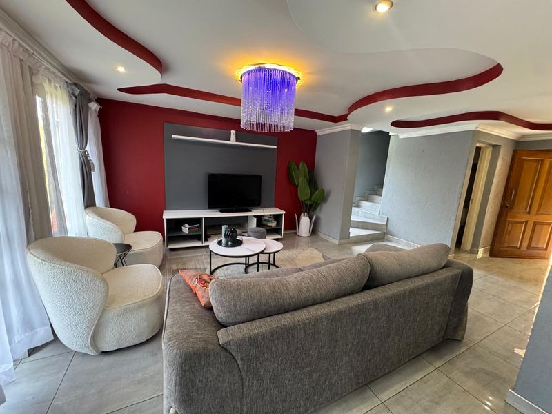A living room featuring a distinctive recessed ceiling with red accents, tiled flooring, and a built-in media wall, leading to an internal staircase.