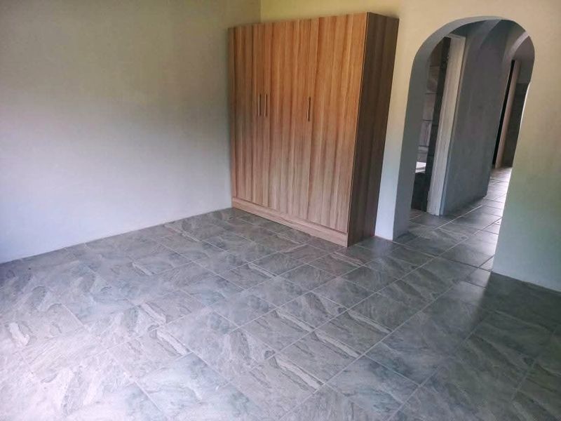 A spacious bedroom featuring neutral tiled flooring, a built-in wooden wardrobe, and an arched entryway leading to the hallway.