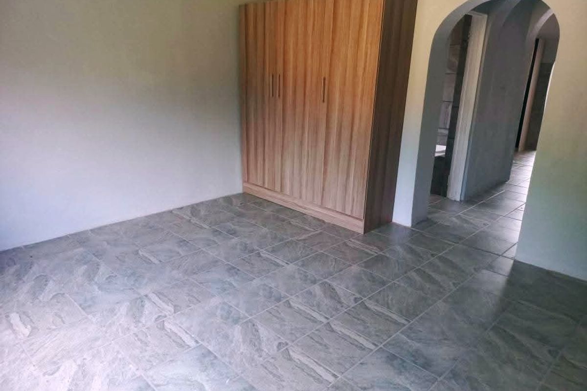A spacious bedroom featuring neutral tiled flooring, a built-in wooden wardrobe, and an arched entryway leading to the hallway.