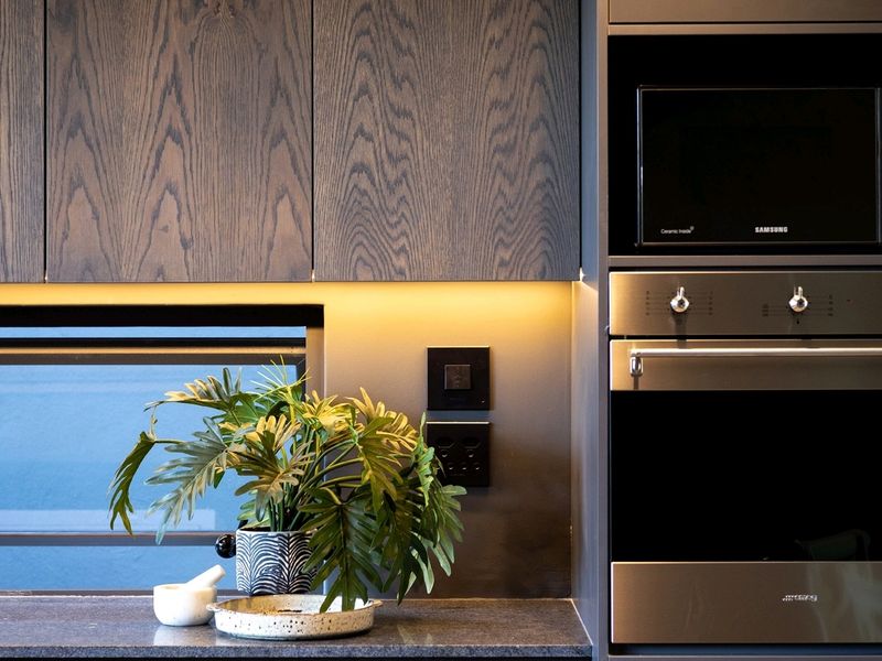 Modern kitchen featuring dark wood cabinetry, stone countertops, integrated appliances including a built-in oven and microwave, and ambient under-cabinet lighting.