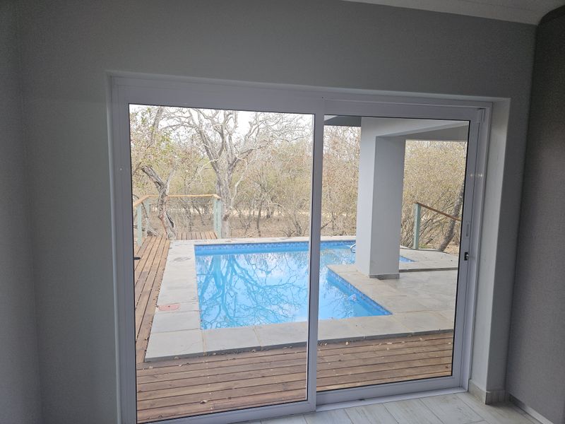 Outdoor pool area featuring timber decking, stone coping, and a covered patio, viewed through a sliding glass door.