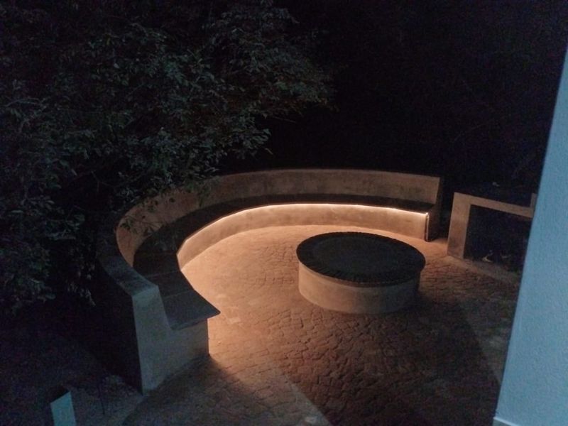 Outdoor patio area featuring a built-in curved concrete bench with integrated LED lighting, a central fire pit, and a masonry barbecue station.