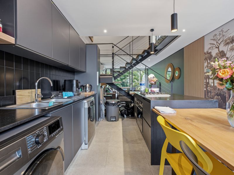 Modern kitchen featuring sleek dark cabinetry, integrated appliances, tiled backsplash, and a seamless transition to an open-plan living area with a striking metal staircase.