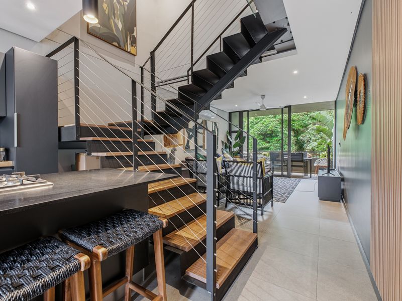 Modern open-plan living area featuring a sleek kitchen, striking timber and steel staircase, and seamless flow to an outdoor balcony through large sliding doors.
