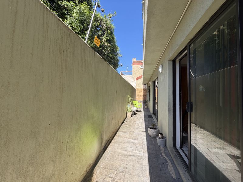 A narrow paved side walkway or patio area featuring a high boundary wall and sliding glass door access to the apartment interior.