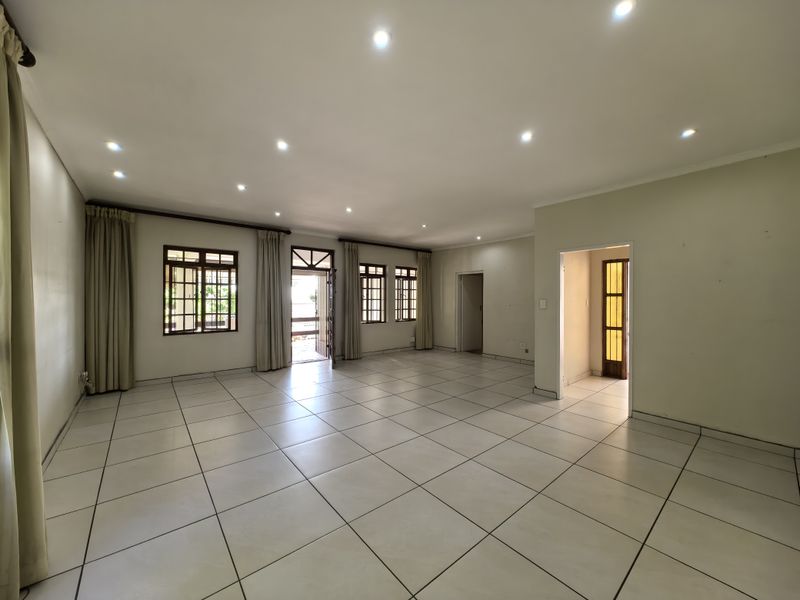 A spacious, empty living area featuring neutral tiled flooring, recessed downlighting, and timber-framed windows and doors leading to an outdoor space.