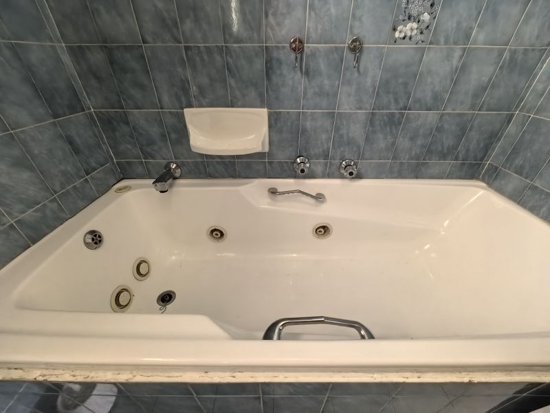 A dated bathroom featuring a built-in jetted bathtub with blue wall tiling and chrome fixtures, requiring minor maintenance to the sealant and grout.