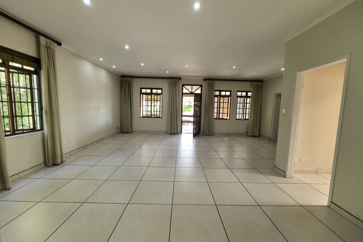 A spacious, empty living area featuring neutral tiled flooring, recessed downlighting, and multiple timber-framed windows providing natural light and access to the exterior.