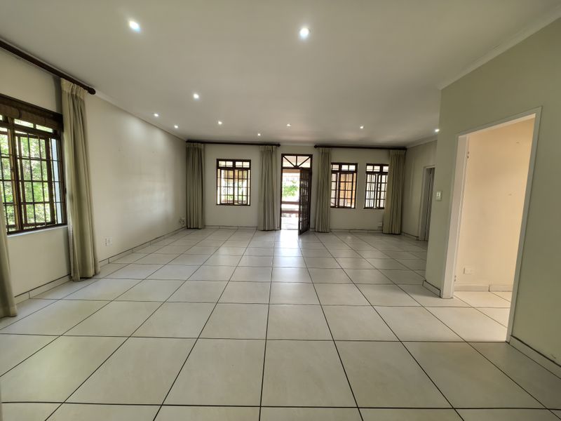 A spacious, empty living area featuring neutral tiled flooring, recessed downlighting, and multiple timber-framed windows providing natural light and access to the exterior.
