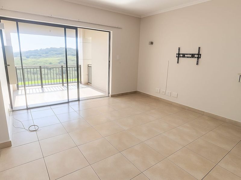 Spacious, empty living area featuring neutral tiled flooring, a wall-mounted TV bracket, and large sliding glass doors opening onto a balcony with scenic views.