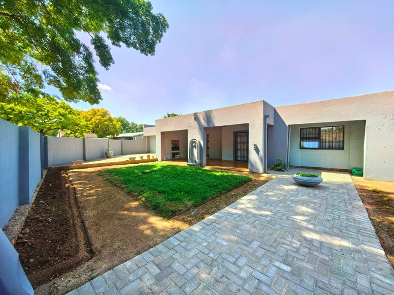 Rear exterior view featuring a paved walkway, a covered patio area, a central lawn, and a perimeter wall with a built-in water feature.