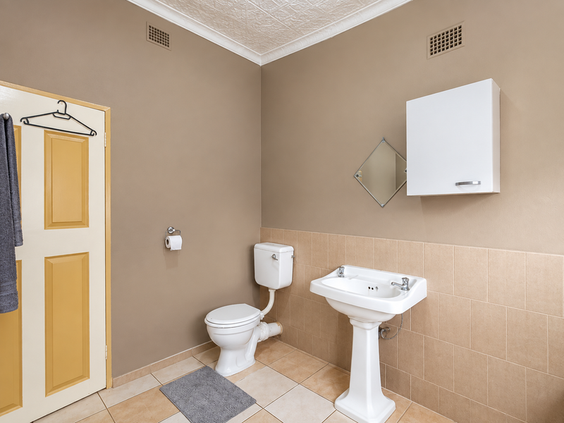 A functional bathroom featuring a pedestal basin, toilet, wall-mounted storage cabinet, and neutral tiled finishes with decorative ceiling detailing.