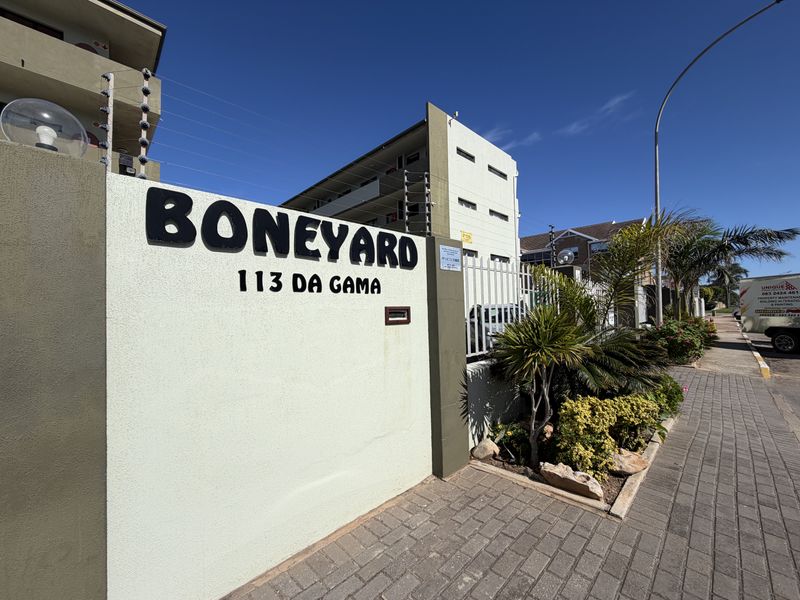 Front exterior view of the apartment complex entrance, featuring a perimeter wall with electric fencing and paved sidewalk access.