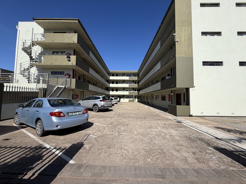 Exterior view of a multi-story apartment complex featuring open-air balcony walkways, concrete staircases, and a paved parking courtyard.