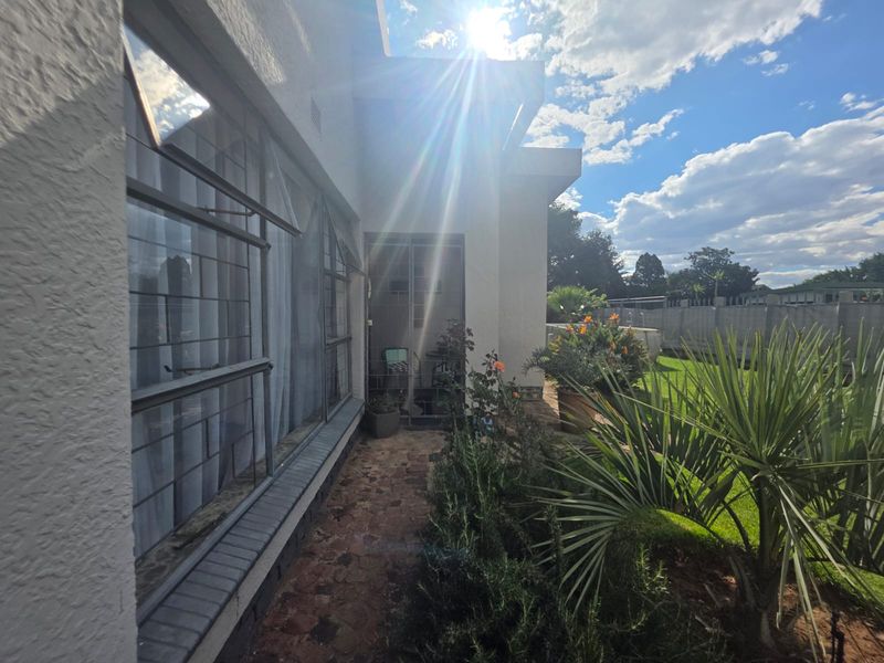 A view of the rear exterior of the house, featuring a paved walkway alongside large steel-framed windows and a well-maintained garden area.