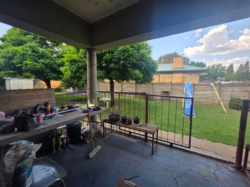 A covered outdoor patio area featuring a concrete floor, metal railing, and a support pillar, overlooking a private backyard garden.