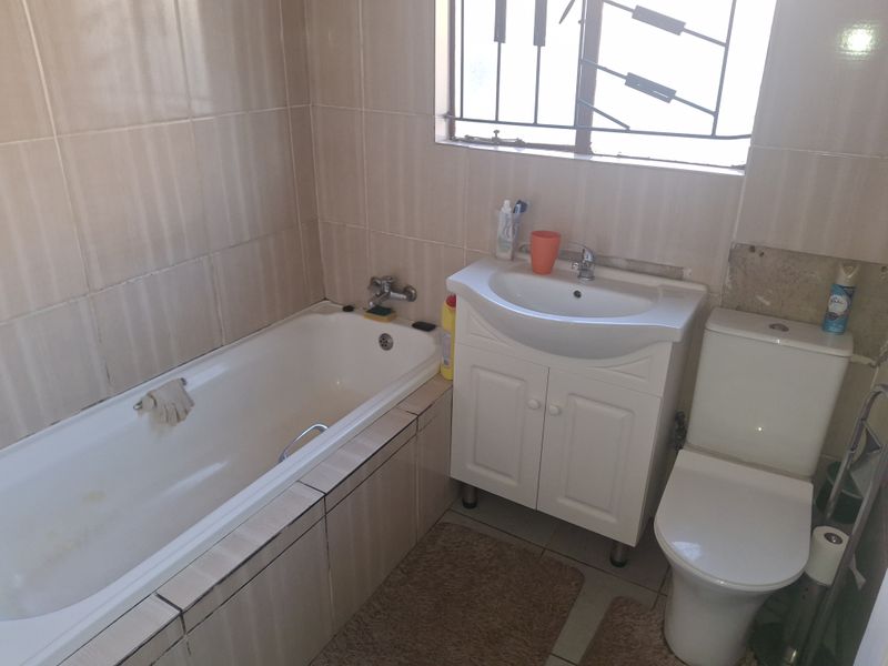 A compact bathroom featuring a bathtub, vanity unit with basin, and toilet, with tiled walls and security bars on the window.
