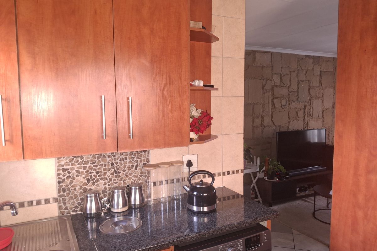 A functional kitchen featuring warm-toned wooden cabinetry, granite countertops, and a tiled backsplash, seamlessly flowing into an adjacent living area with a stone feature wall.