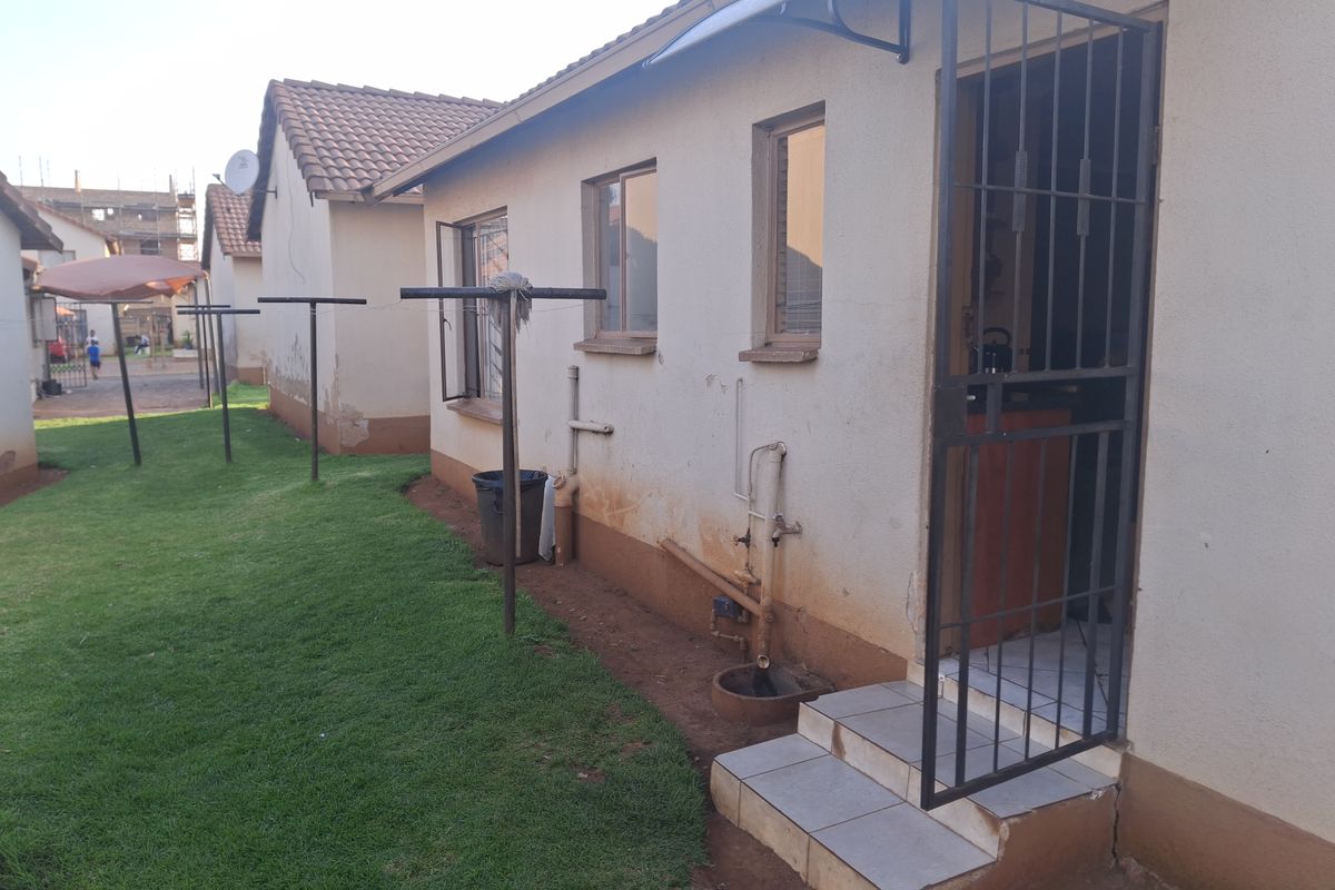 Rear exterior view of the property featuring a security gate, tiled entrance steps, and external plumbing fixtures set against a small lawn area.