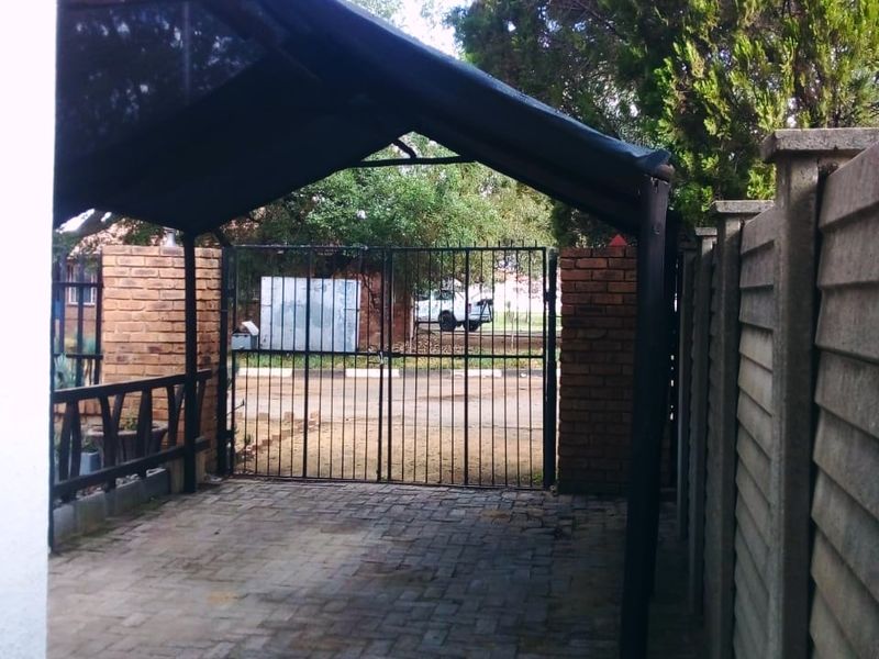 A paved outdoor parking area featuring a metal carport structure, enclosed by brick and precast concrete boundary walls with a secure metal gate.