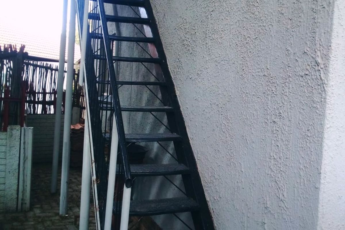 An exterior view of a metal staircase providing access to an upper level, set against a textured exterior wall with paved ground below.