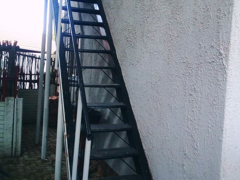An exterior view of a metal staircase providing access to an upper level, set against a textured exterior wall with paved ground below.