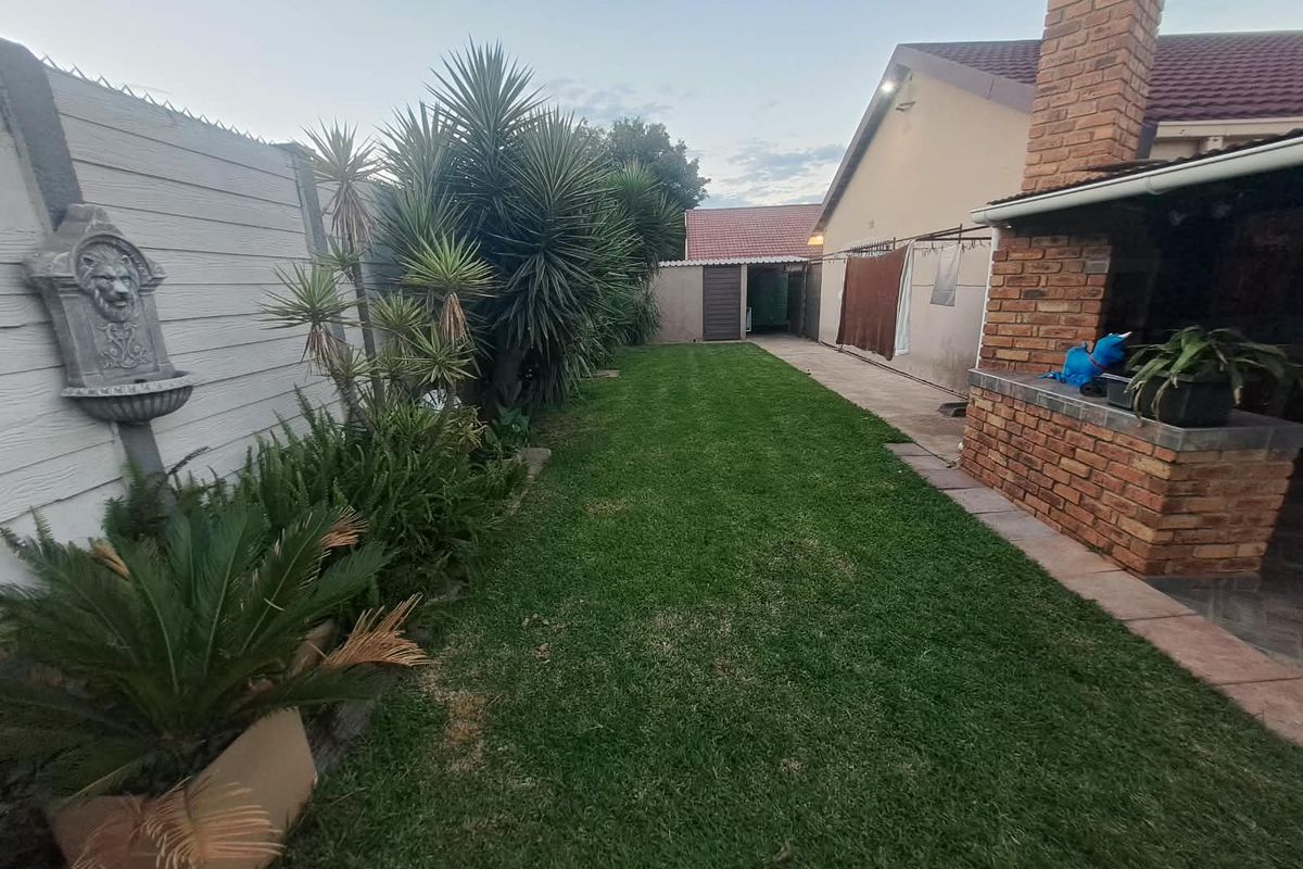 A private rear garden featuring a well-maintained lawn, a brick built-in braai area, and a paved walkway along the side of the house.