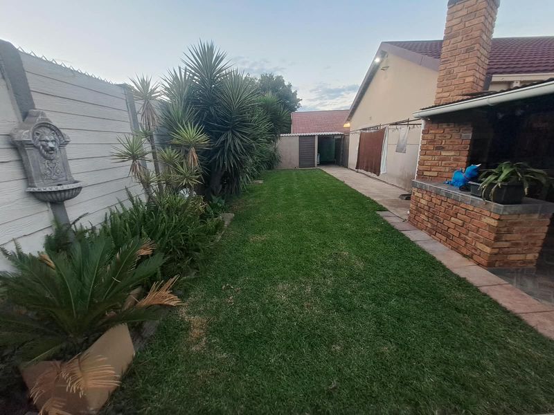 A private rear garden featuring a well-maintained lawn, a brick built-in braai area, and a paved walkway along the side of the house.