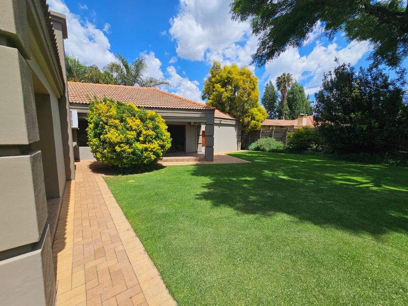 A well-maintained rear garden featuring a lush green lawn, a paved walkway, and a covered patio area attached to the main house structure.