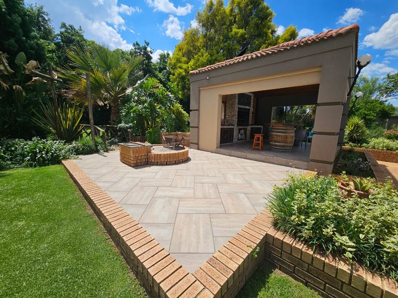 A well-maintained covered patio featuring a built-in braai, tiled flooring, and an adjacent outdoor fire pit area set within a landscaped garden.