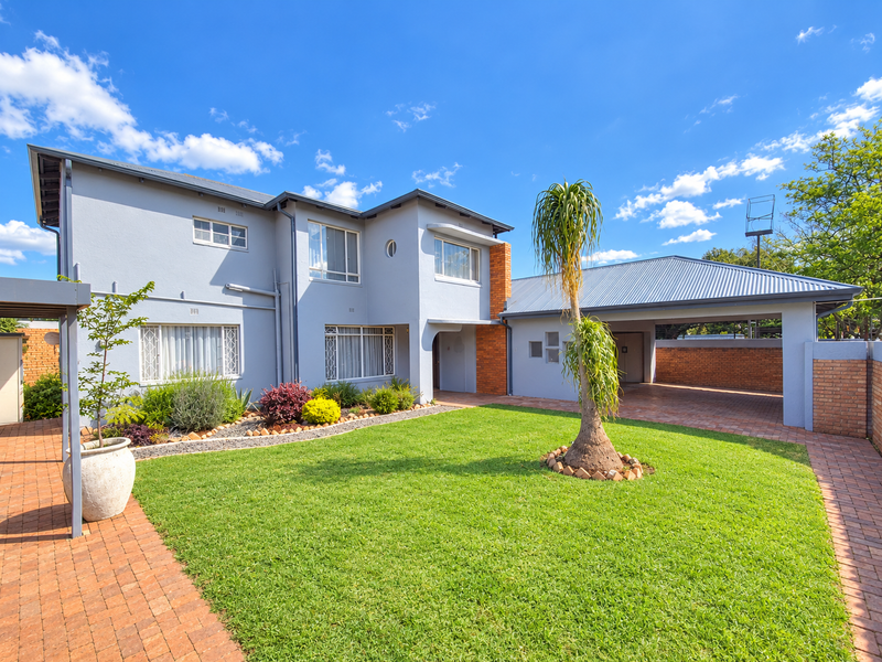 A well-maintained double-story house featuring a paved brick driveway, attached carport, and a neatly landscaped front garden under a clear blue sky.