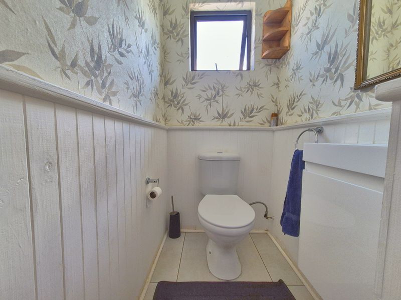 A compact bathroom featuring wainscoting, patterned wallpaper, a white toilet, and a vanity unit with tiled flooring.