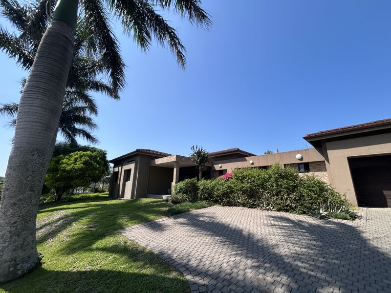 A well-maintained single-story house exterior featuring a paved driveway, covered entrance, and established garden landscaping under a clear blue sky.