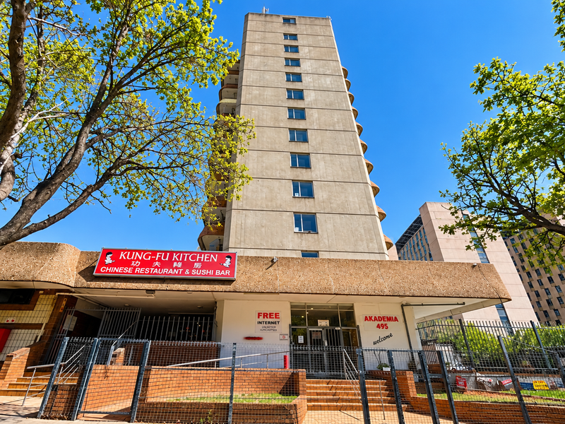 Front exterior view of a multi-story apartment building featuring a concrete facade, ground-level commercial space, and secure perimeter fencing.