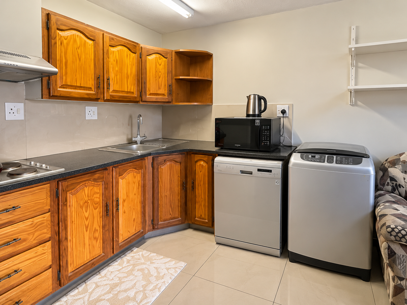 A functional kitchen featuring wooden cabinetry, a dark laminate countertop, stainless steel sink, built-in hob with extractor fan, and tiled flooring.