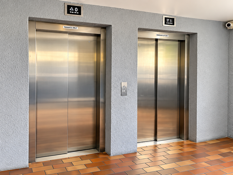A clean and well-maintained apartment building elevator lobby featuring dual stainless steel lift doors, textured walls, and durable tiled flooring.