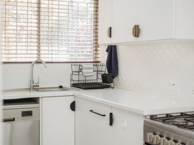 A bright, modern kitchen featuring white cabinetry, a stylish scalloped tile backsplash, stainless steel sink, and integrated dishwasher.