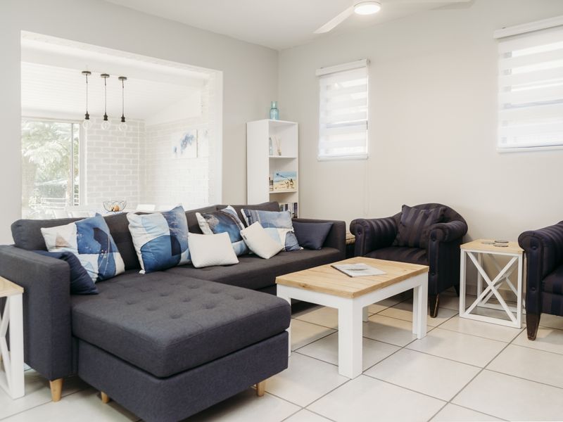 A bright and well-maintained living room featuring neutral tiled flooring, modern ceiling fan, and roller blinds, with an open transition to an adjacent space.