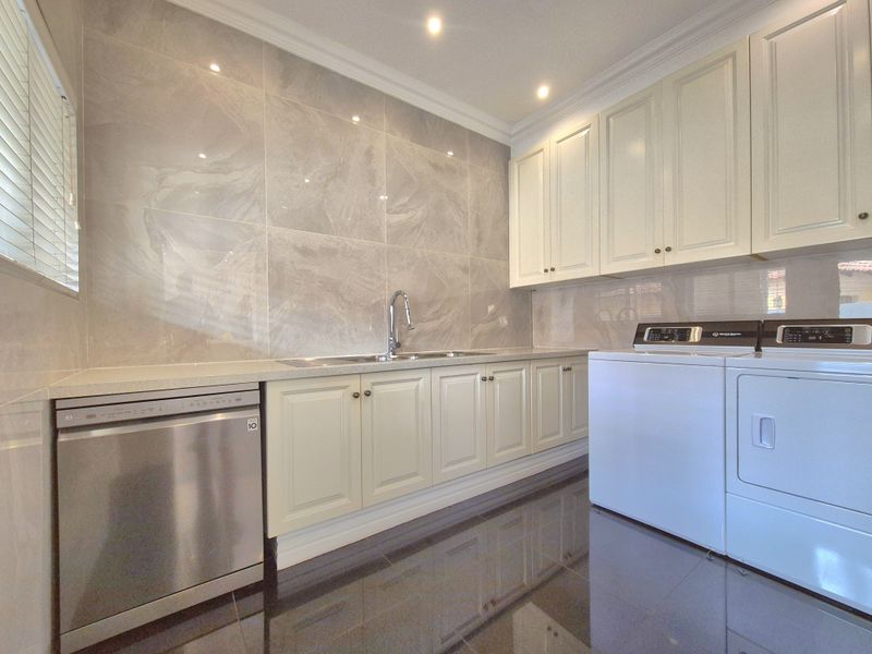 A well-appointed laundry room featuring sleek cabinetry, stone benchtops, a stainless steel sink, and modern tiled finishes throughout.