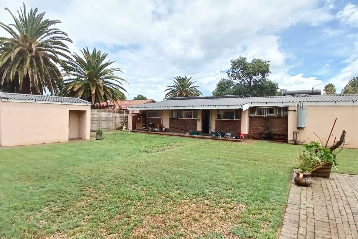 Rear view of the property featuring a spacious lawn, paved walkway, detached outbuilding, and brick facade with a metal roof.