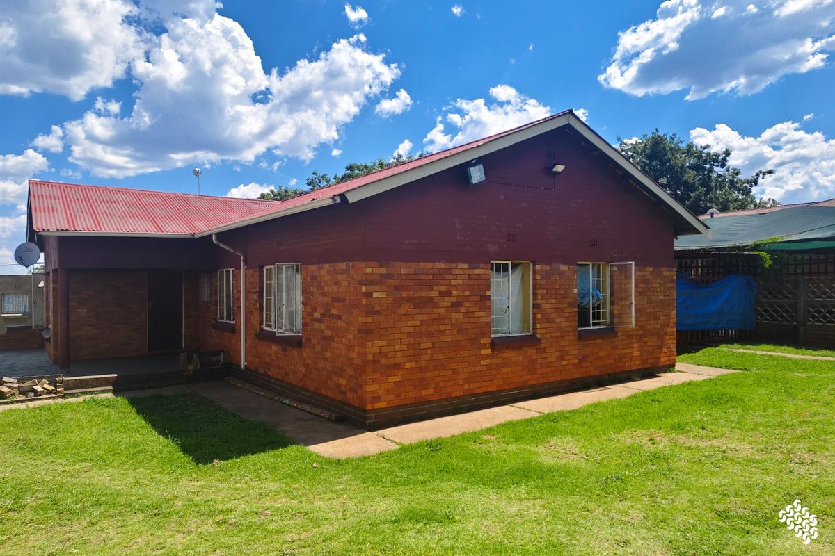 Rear view of a single-story brick house featuring a red corrugated metal roof, security bars on windows, and surrounding lawn area.